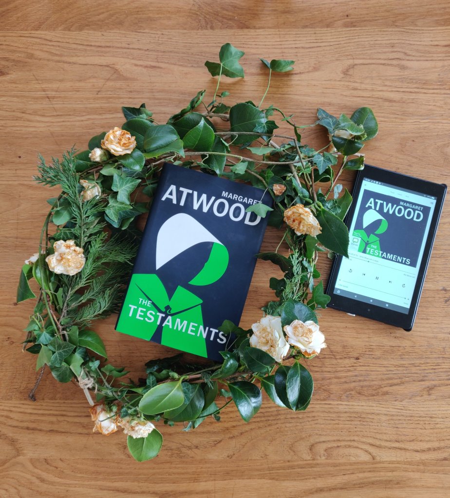 Image of a hardback copy of the Treatments, by Margaret Atwood, in a wreath and a kindle places next to it with the audio version of the same book. 