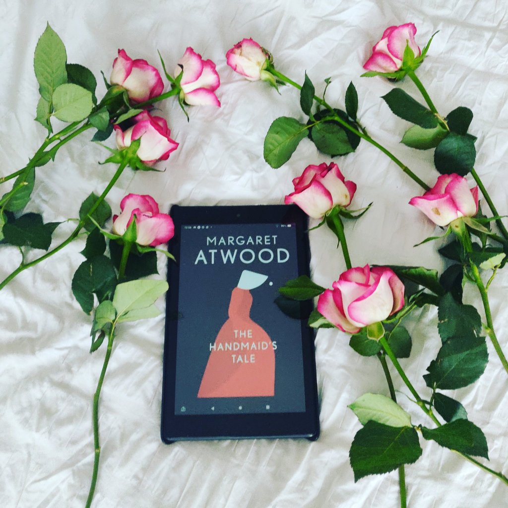 Image of a kindle devise with the Handmaid's Tale book cover, surrounded by pink roses on a bed. 