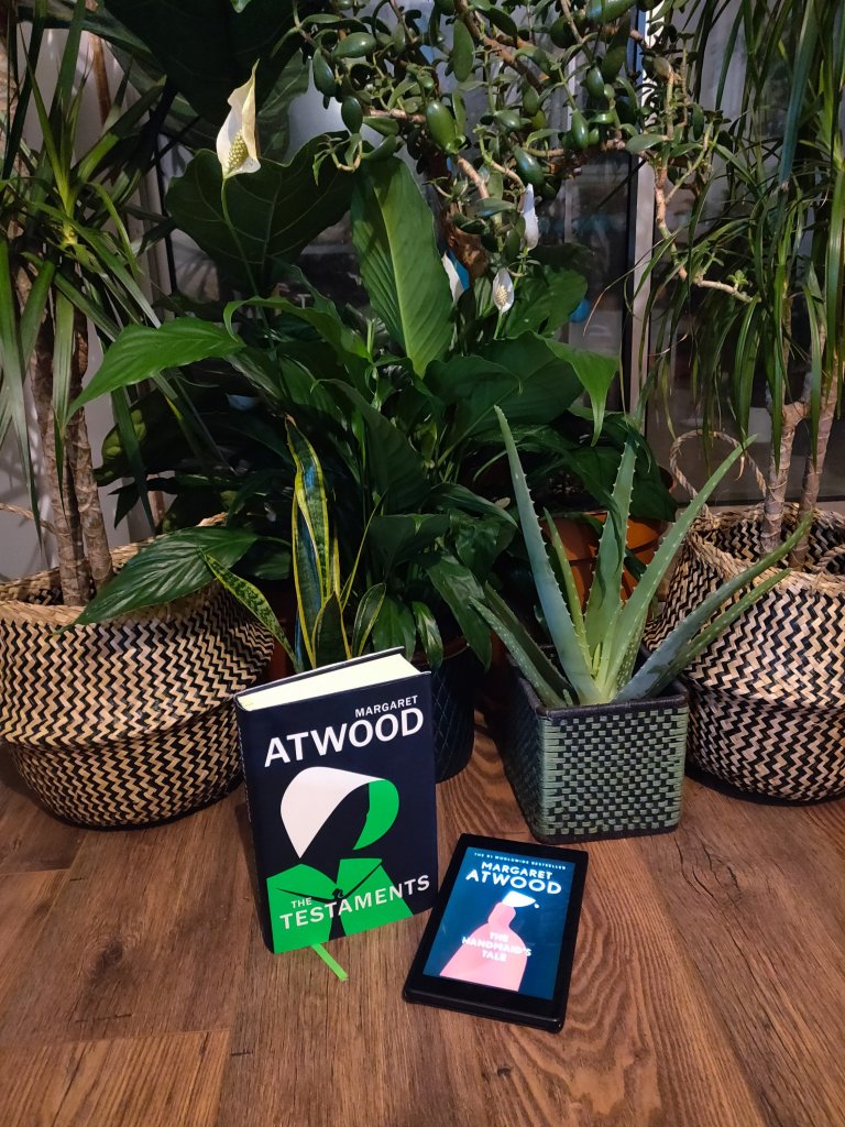 Image of the hardback copy of the Testaments and a kindle displaying the cover of The Handmaid's Tale, in front of some house plants.
