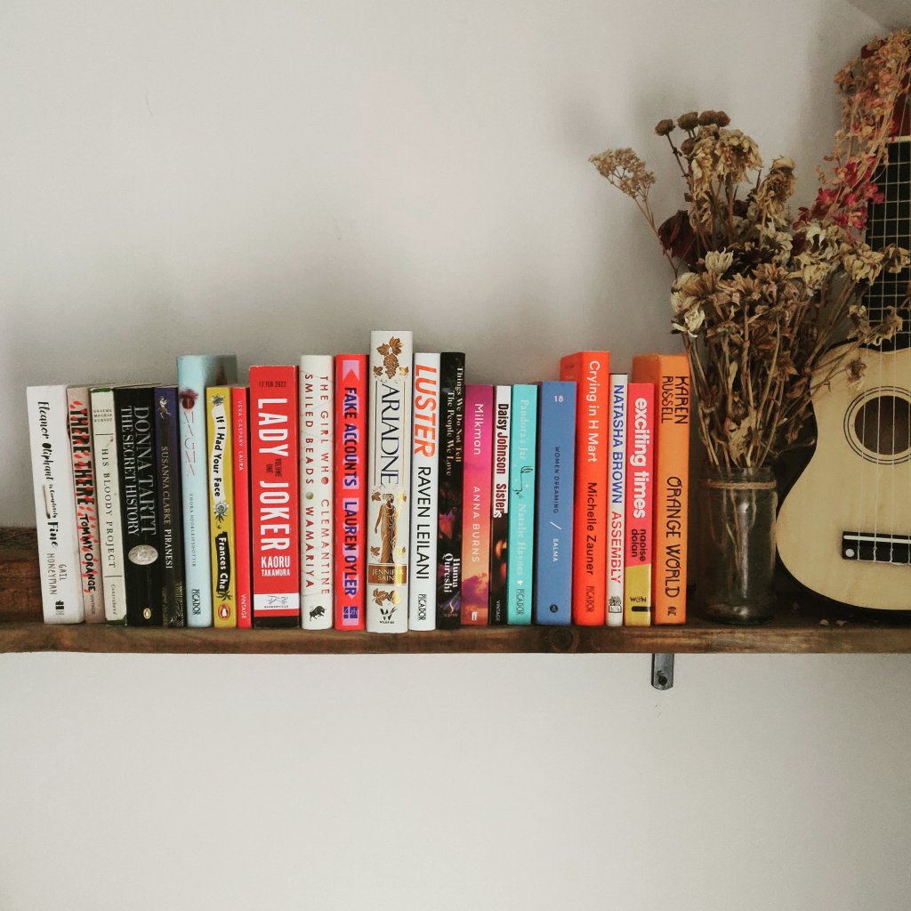 A wooden shelf with twenty-two books I'd like to read this year. There is also a vase of dried flowers and a ukulele at the end of the shelf.  