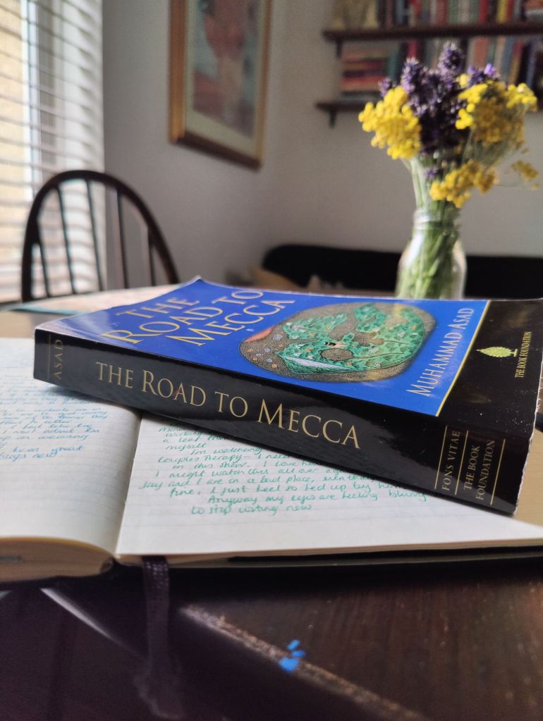A copy of The Road To Mecca by Muhammad Asad on top of an open journal with a glass bottle with flowers in it in the background. 